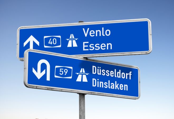 Zwei blaue Autobahnschilder mit weißen Richtungspfeilen. Auf einem steht „40 Venlo Essen“, auf dem anderen „59 Dinslaken Duisburg“. Die Schilder symbolisieren die Lage im westlichen Ruhrgebiet.
