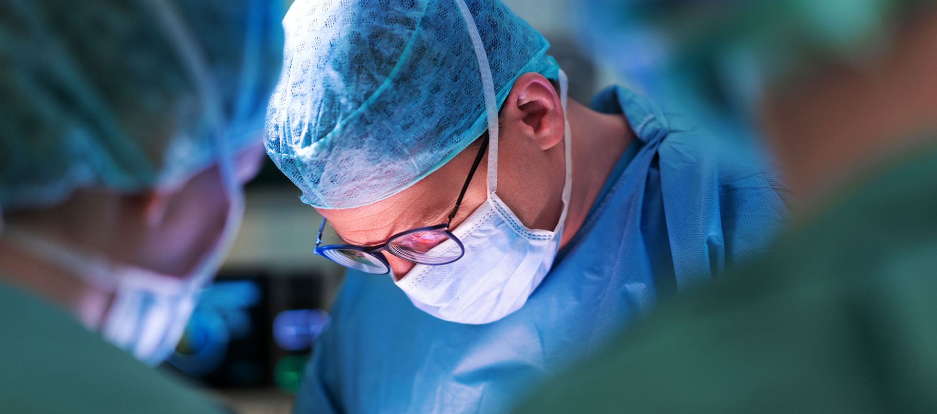 Ein Chirurg im OP, konzentriert bei der Durchführung eines Eingriffs. Er trägt vollständige Schutzkleidung mit OP-Kittel, Haube, Handschuhe und Mundschutz.