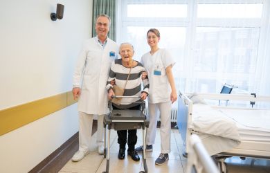 Martha Henze, 110 Jahre alt, steht im Krankenzimmer mit Chefarzt Dr. Dietmar Kumm und Gesundheits- und Krankenpflegerin Sally nach ihrer erfolgreichen Hüftoperation. Dr. Kumm und Sally stützen Martha Henze.
