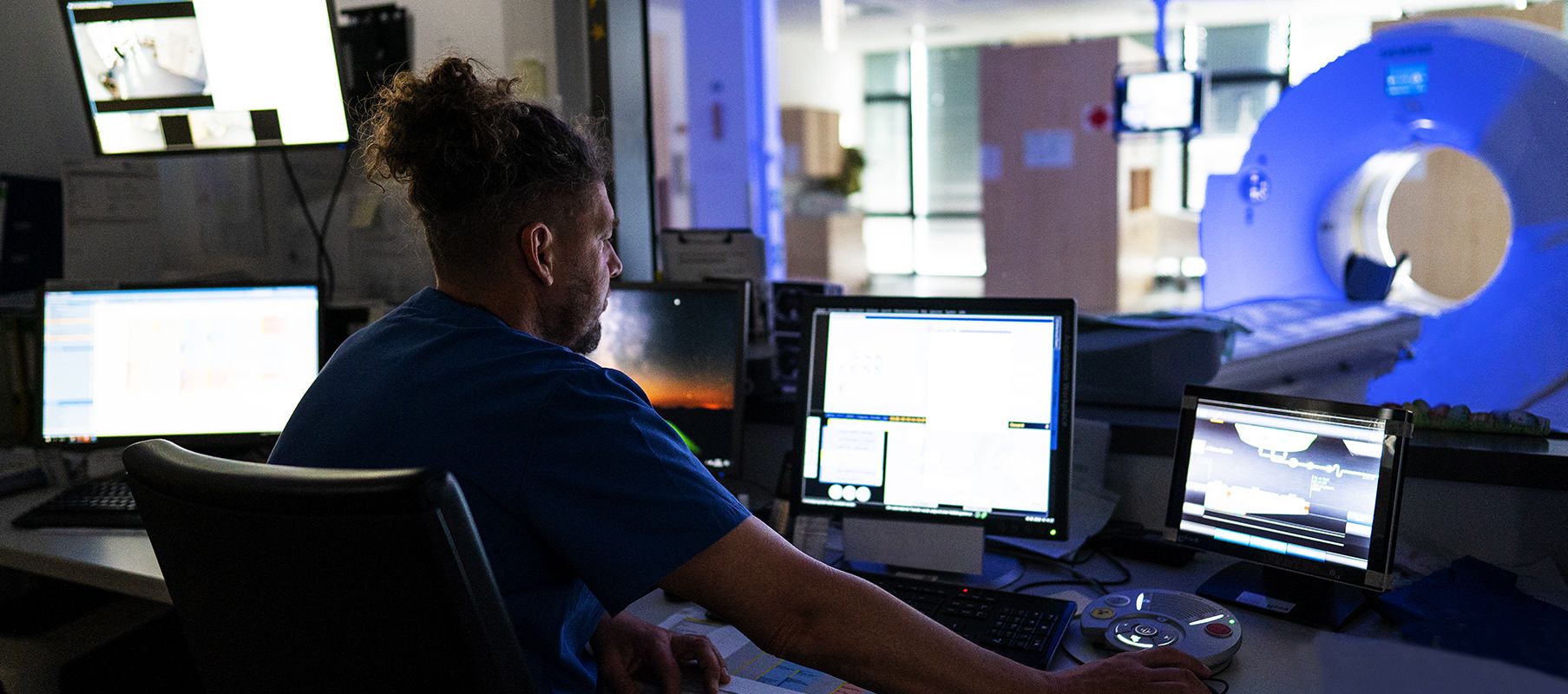 Mitarbeiter sitzt an einem Schreibtisch in der Radiologie-Leitstelle, mit Blick auf den benachbarten CT-Raum. Auf dem Tisch sind Monitore und Geräte sichtbar.