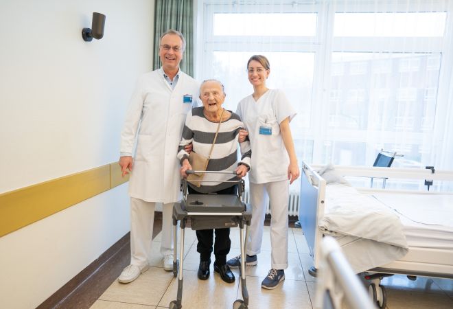 Martha Henze, 110 Jahre alt, steht im Krankenzimmer mit Chefarzt Dr. Dietmar Kumm und Gesundheits- und Krankenpflegerin Sally nach ihrer erfolgreichen Hüftoperation. Dr. Kumm und Sally stützen Martha Henze.