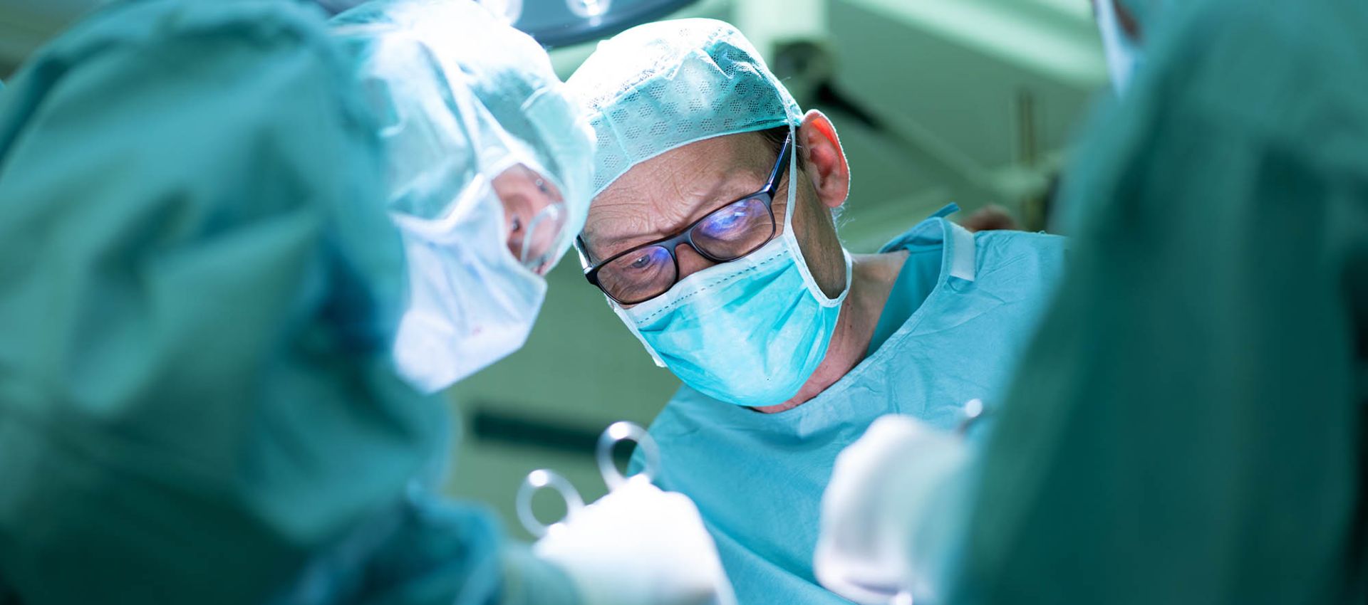 Ein Chirurg im Operationssaal erhält Unterstützung von zwei assistierenden Personen in Schutzkleidung, die ihm präzise Instrumente reichen.