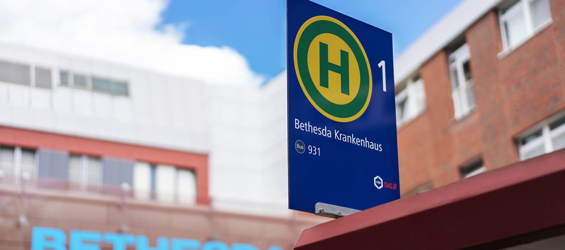 Vorne das Haltestellenschild des Bethesda Krankenhauses, im Hintergrund der Schriftzug ‚Bethesda‘ über der Einfahrt zur Notaufnahme.